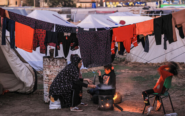 A woman sits by food cooking on a fire as children play nearby outside one of the tents housing Palestinians displaced by the conflict in Gaza between Israel and Palestinian terror group Hamas movement, in Rafah in the southern Gaza Strip on December 18, 2023. (Mahmud Hams / AFP)
